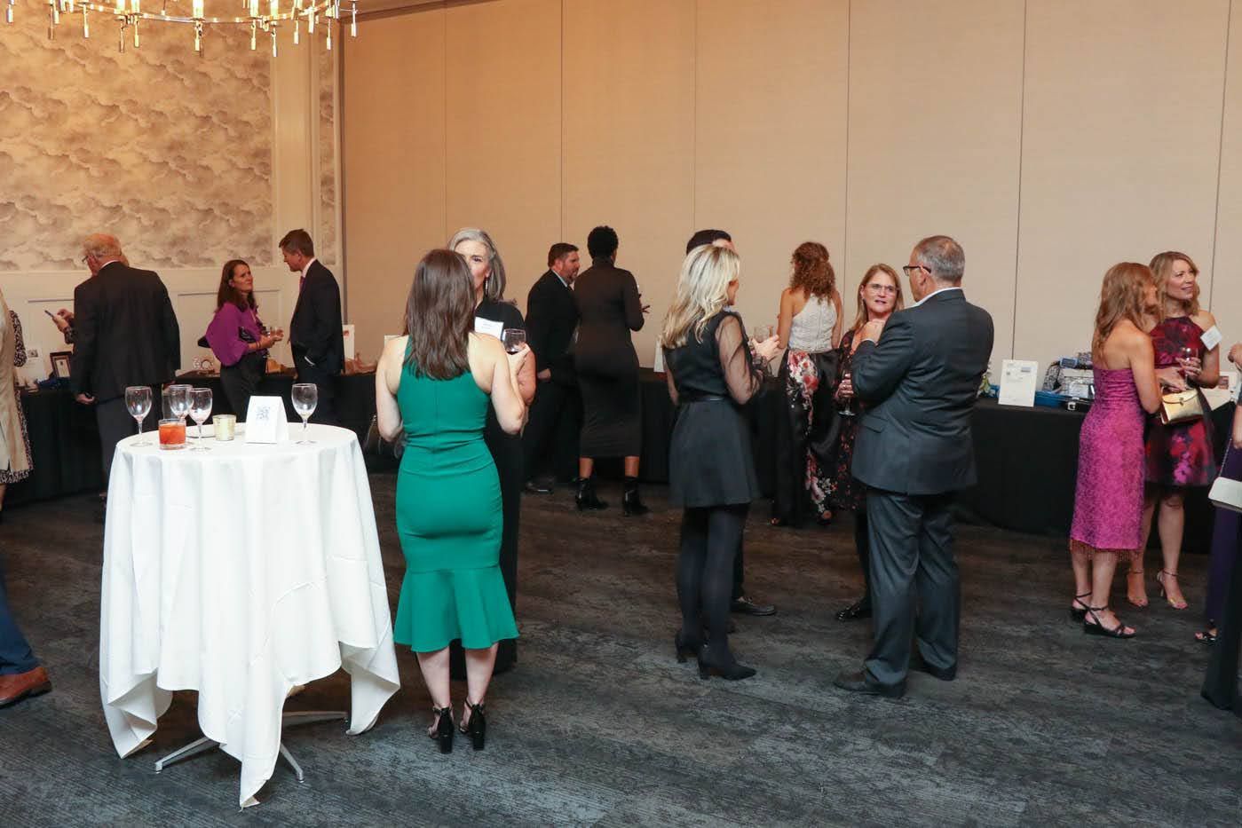 People mingling at an event, some wearing formal attire. A table with a white tablecloth is in the foreground.