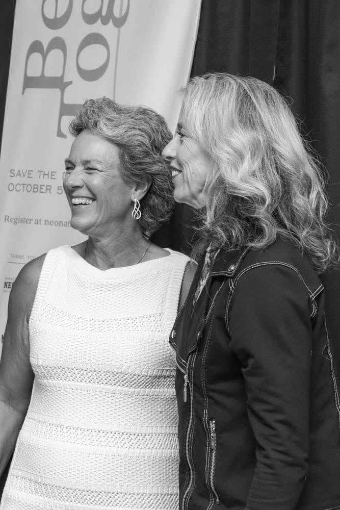 Two smiling women pose together outdoors; one in a white dress, the other in a jacket.