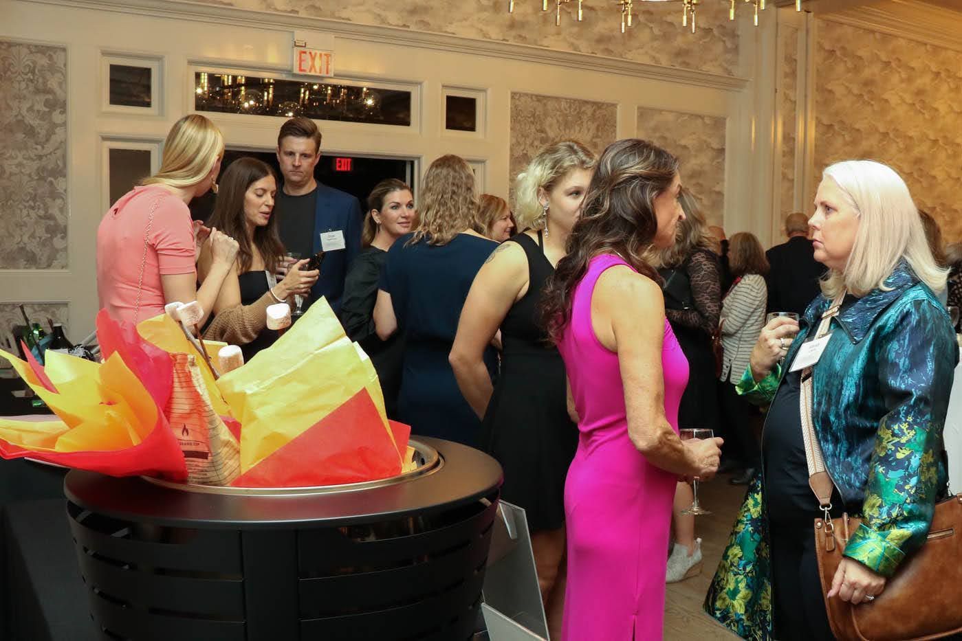 People at a social gathering, talking near a decorative fire pit. Formal attire, neutral wall with exit sign.