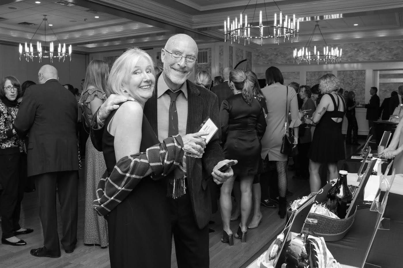 Couple smiling, embracing at a formal event, checkered scarf, many guests and chandeliers in the background.