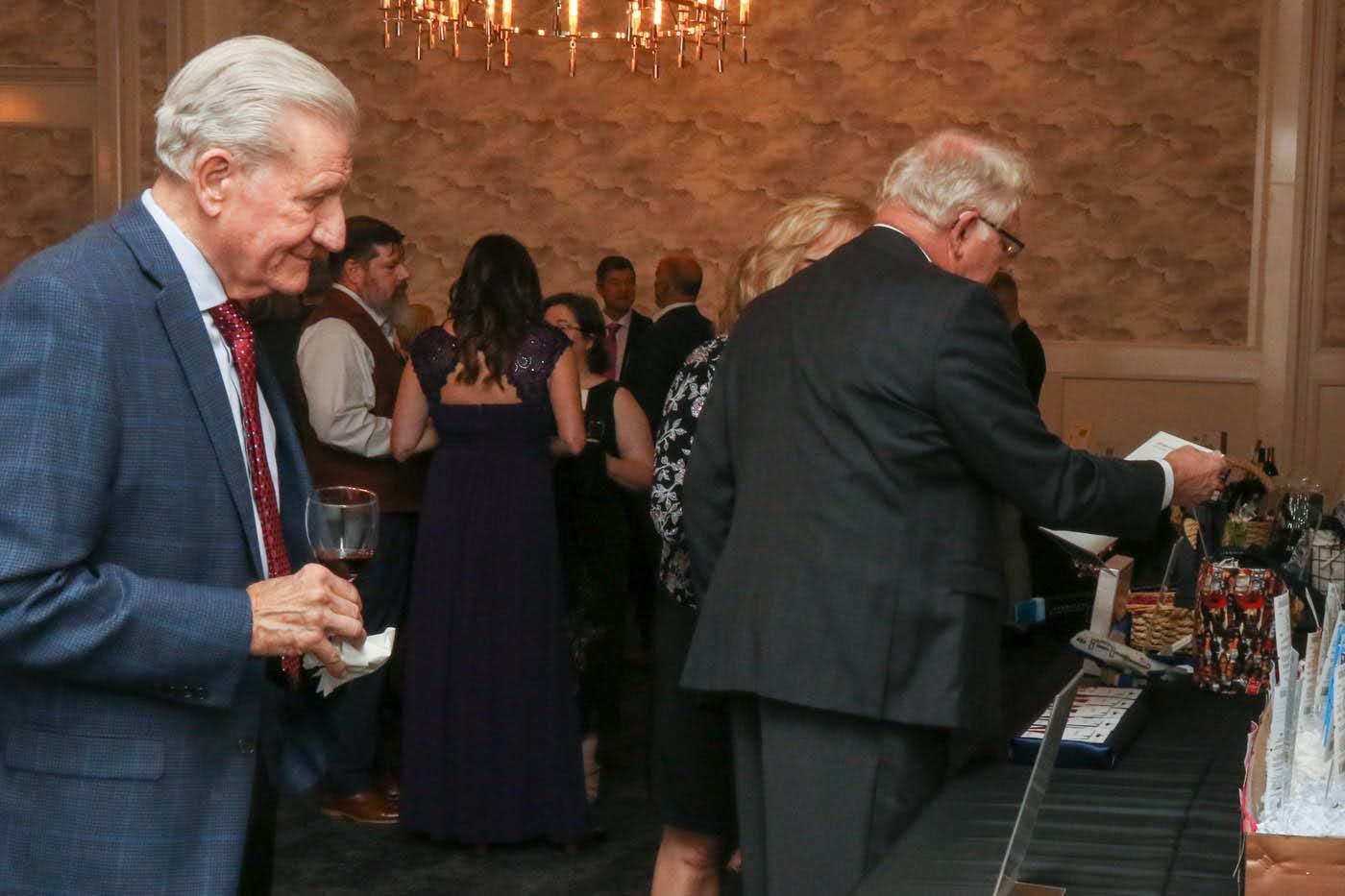 People at an event, with one holding wine and another looking at items on a table. Dim lighting.