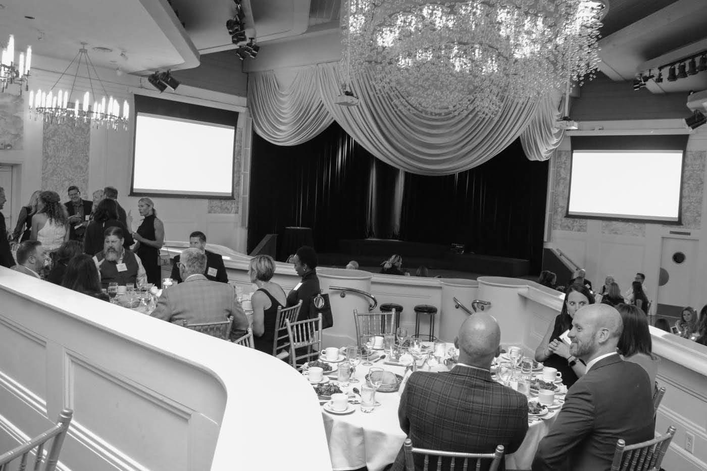 Guests seated at tables in a decorated banquet hall with a stage, screens, and chandelier.