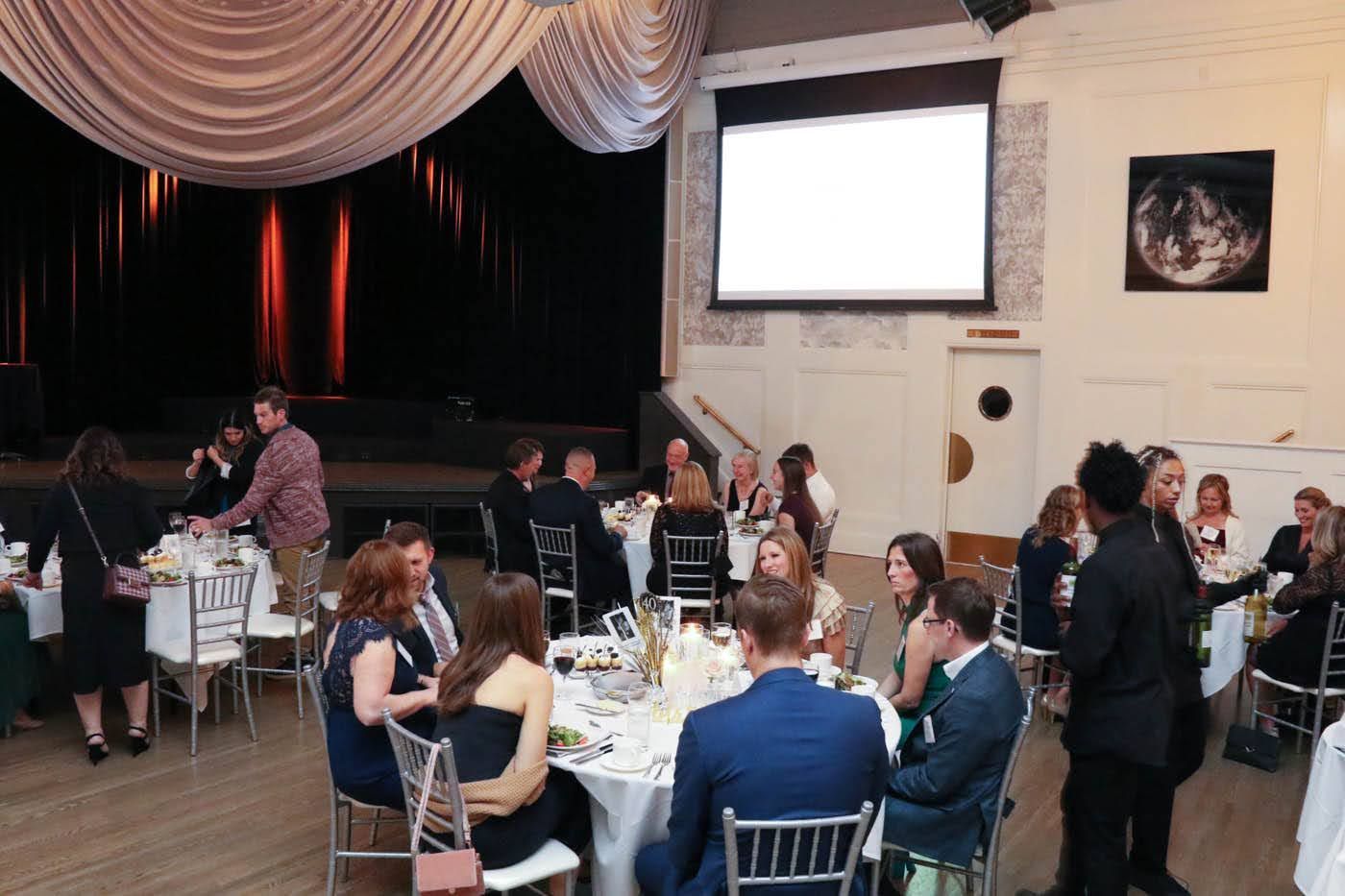 Formal event with guests seated at round tables. A projection screen and stage are visible.
