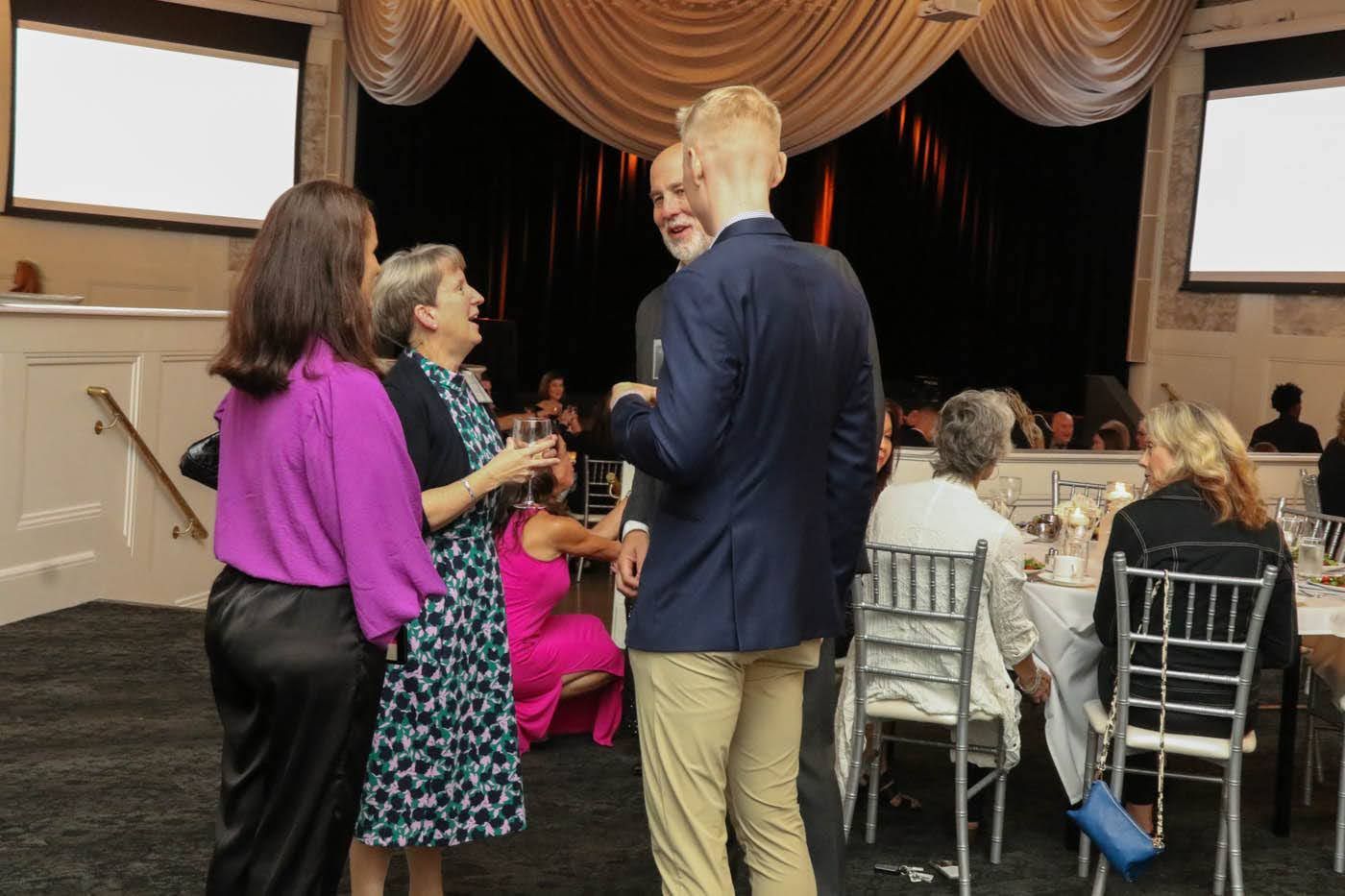 People conversing at an event, some holding drinks. Elegant dining room setting with tables and decorations.
