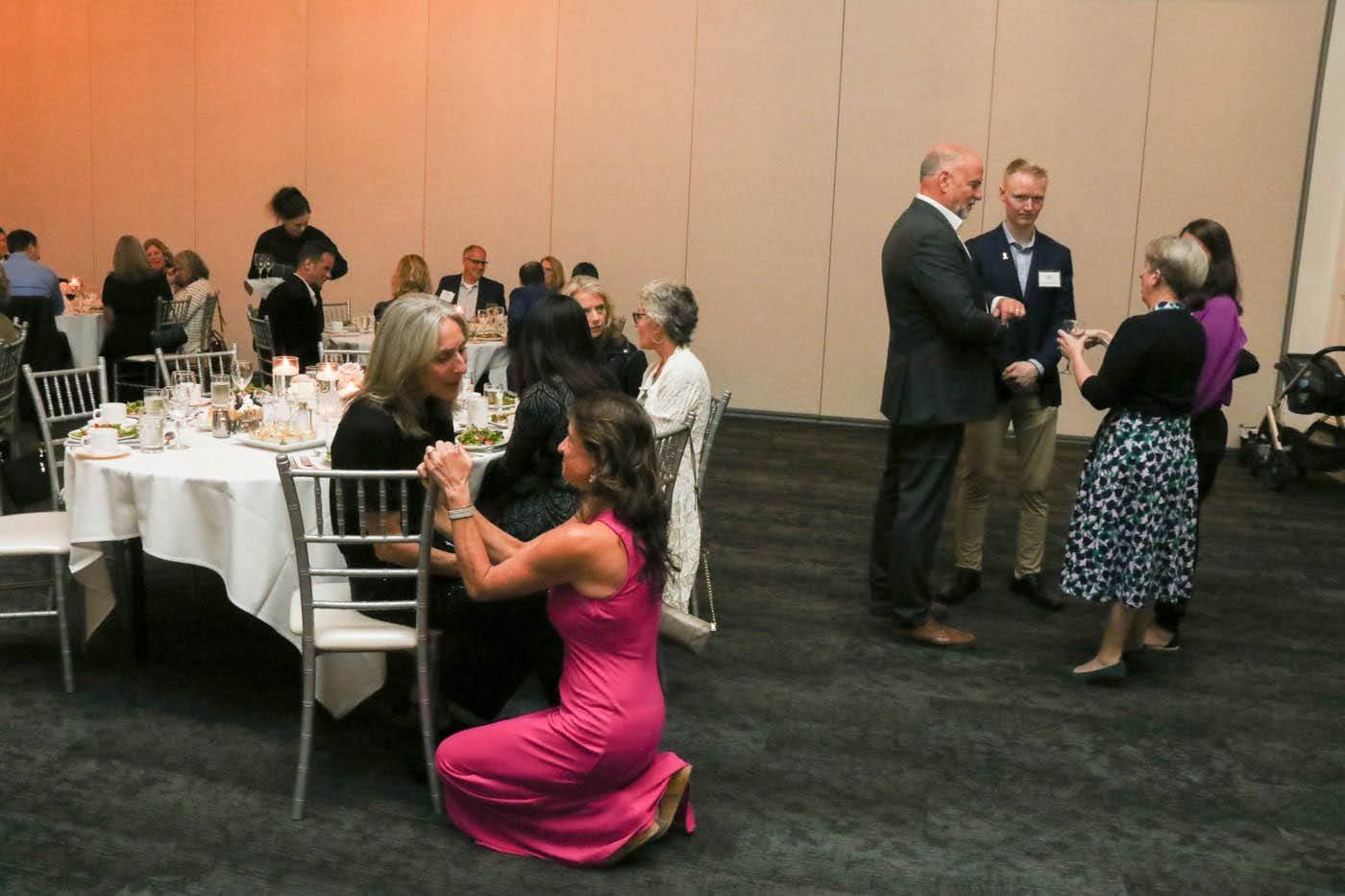 People interacting at a well-lit event. Woman kneels, speaking to another woman seated near a table. Others converse nearby.