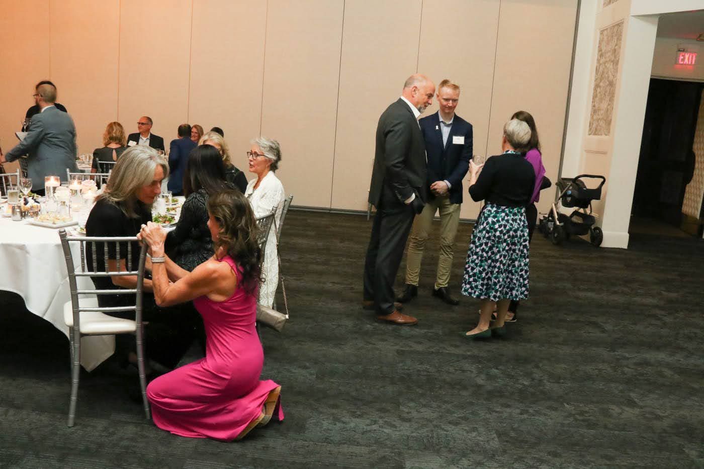 People in a social setting; some are standing, conversing, while others are seated at tables in a brightly lit banquet hall.