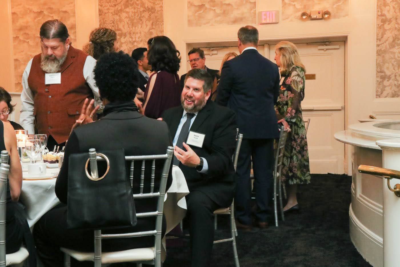 People at an event seated at tables, some talking and standing, in a room with patterned walls and a bar.