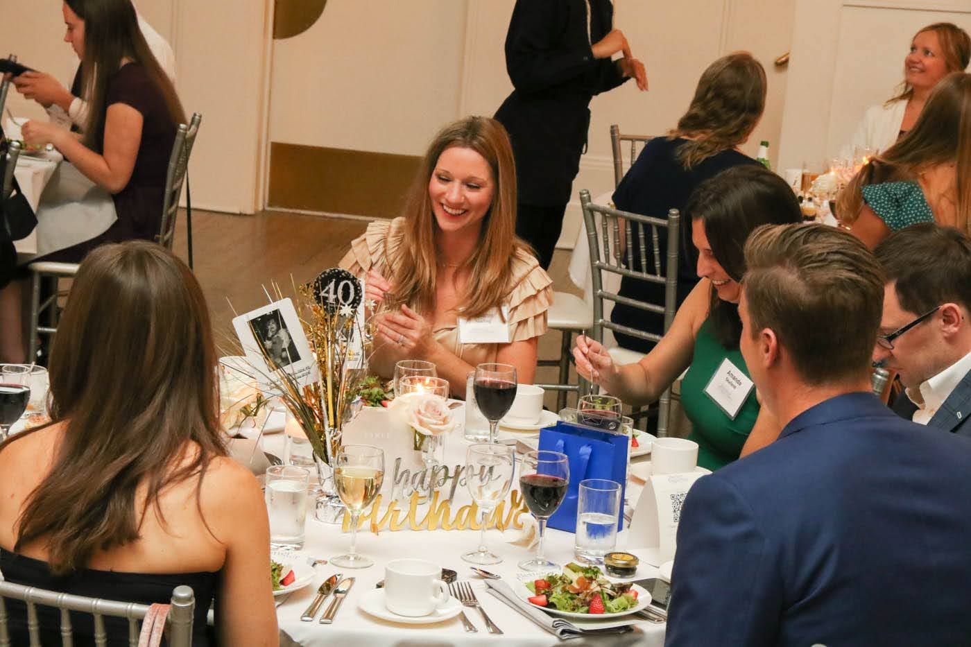 People at a formal dinner, smiling and talking. Table setting with centerpiece and food.