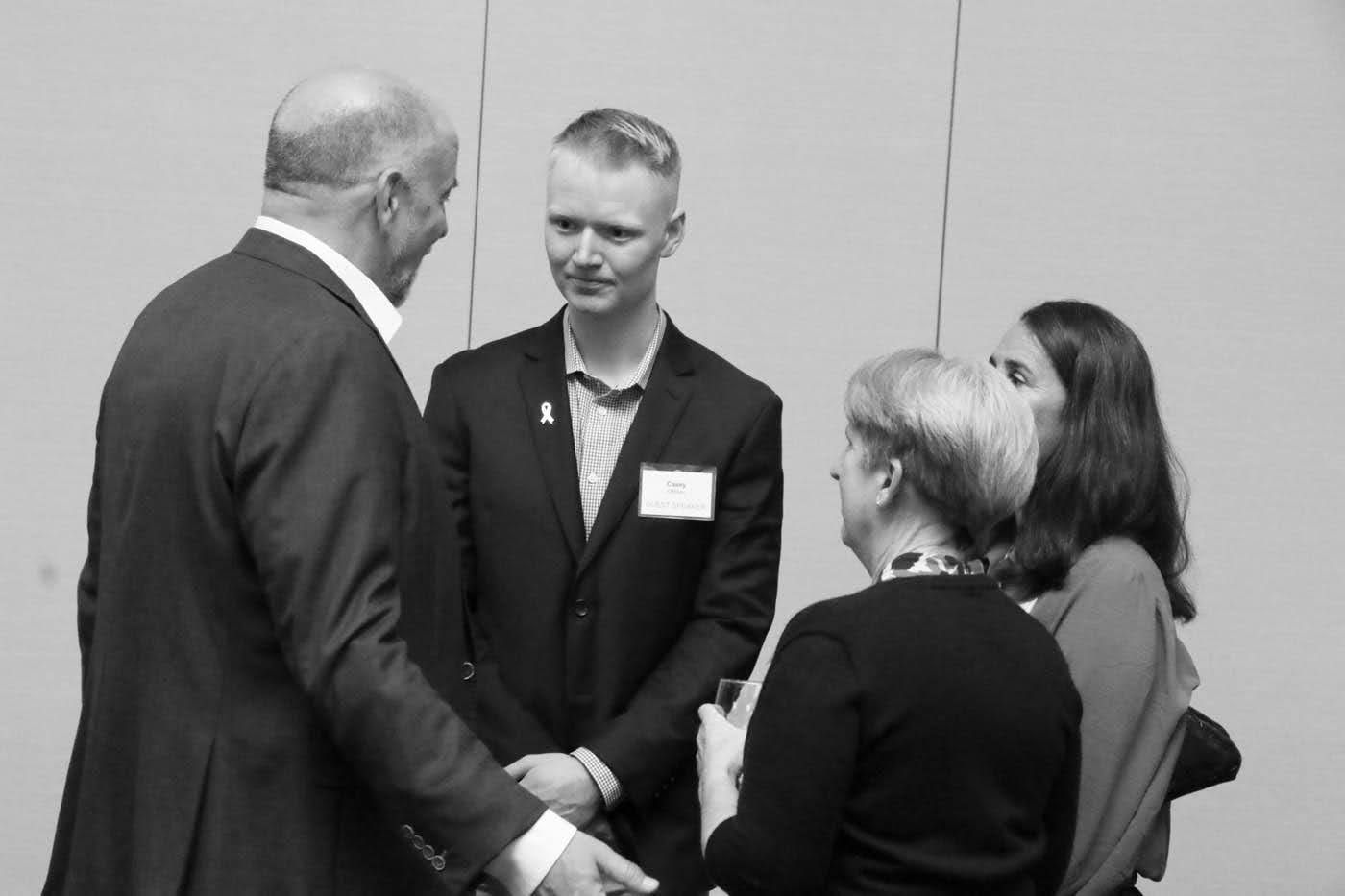 Four people in a room are in a conversation. One man is shaking hands. All are wearing professional attire.
