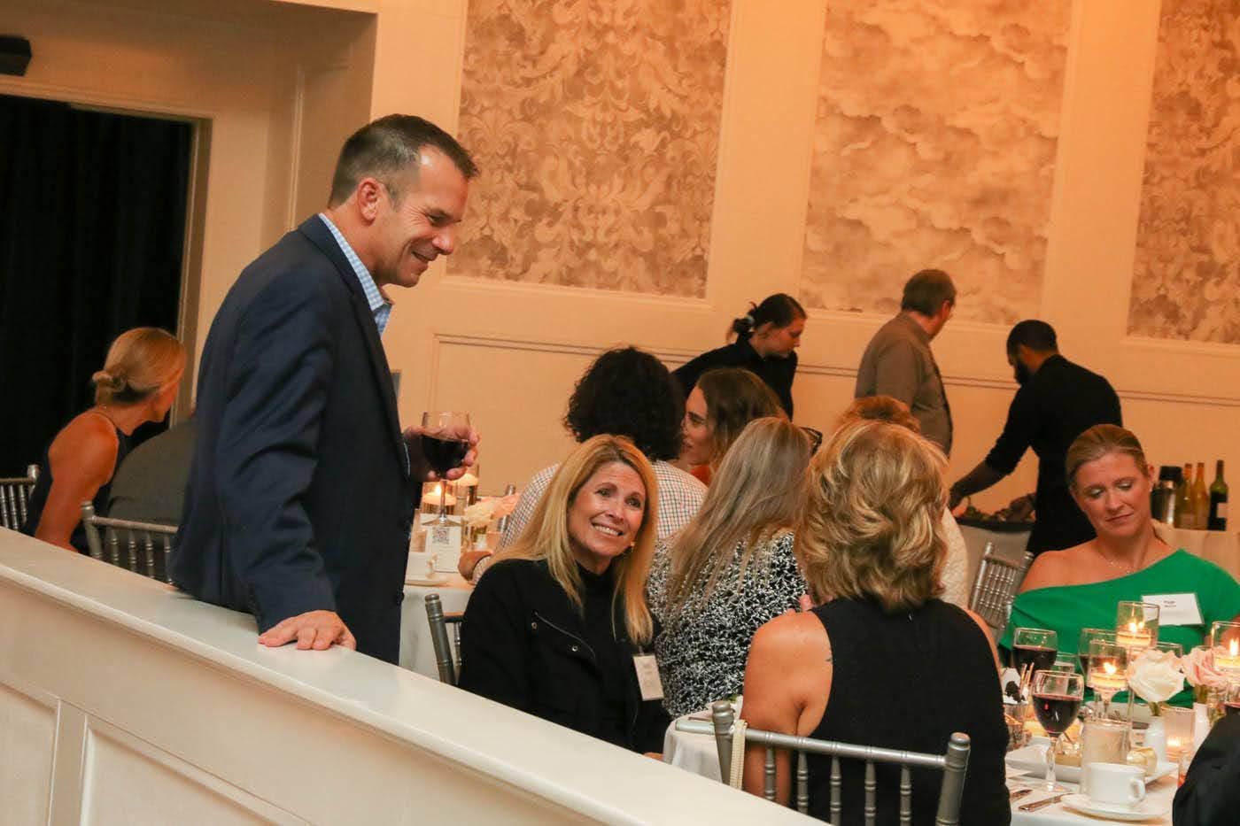 Man in suit chats with seated women at a dinner, lit room, formal event.