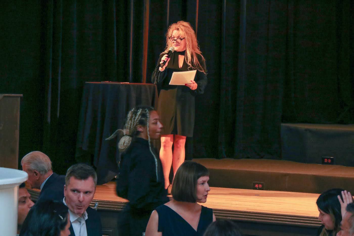 Woman speaking at a formal event on a stage, holding a microphone and papers. Audience members watch.