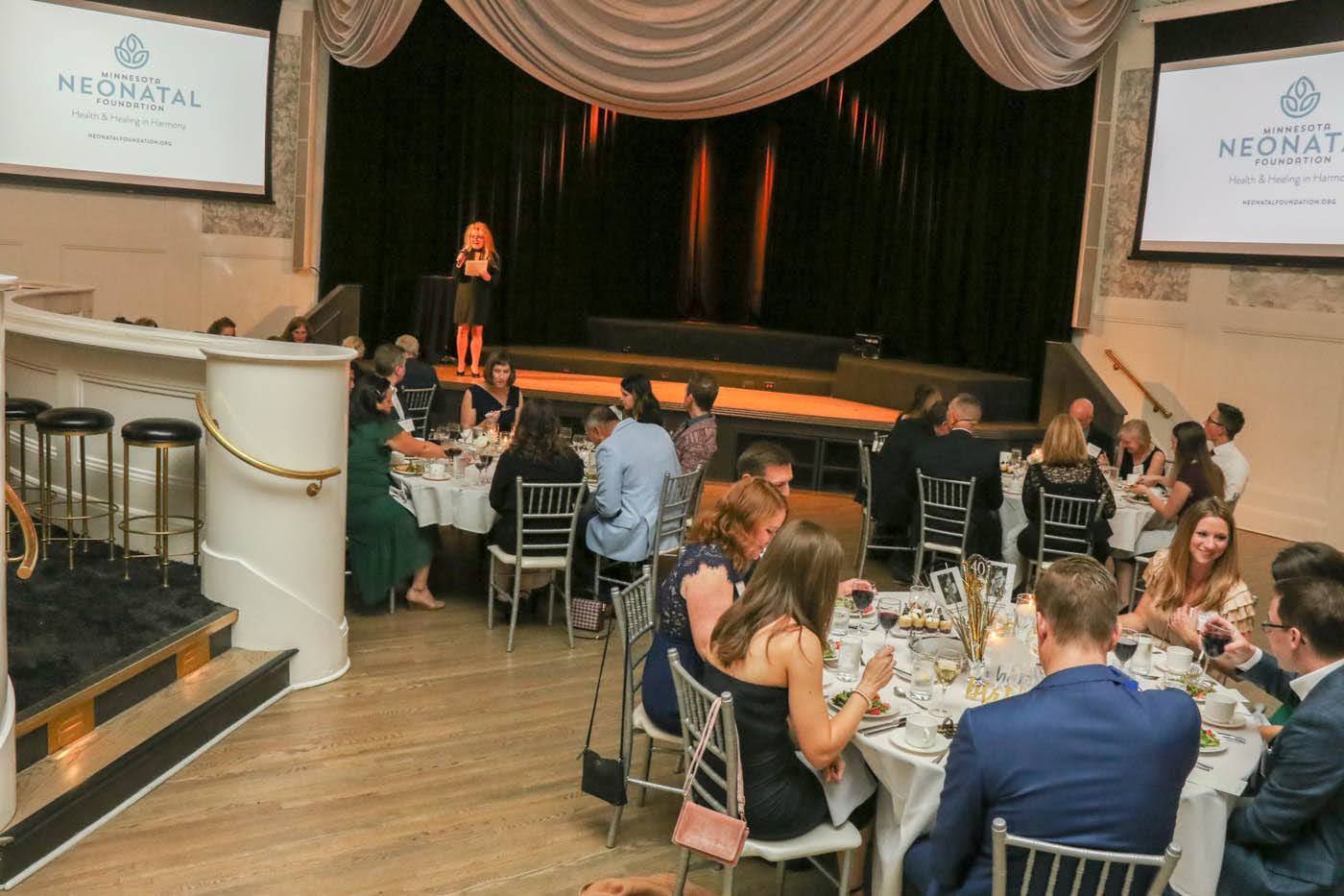 People seated at tables during an event, with a speaker on a stage in the background.