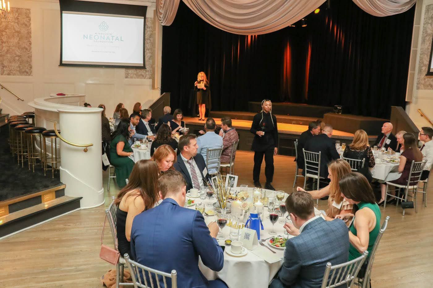 People seated at round tables in a banquet hall, watching a speaker on stage.