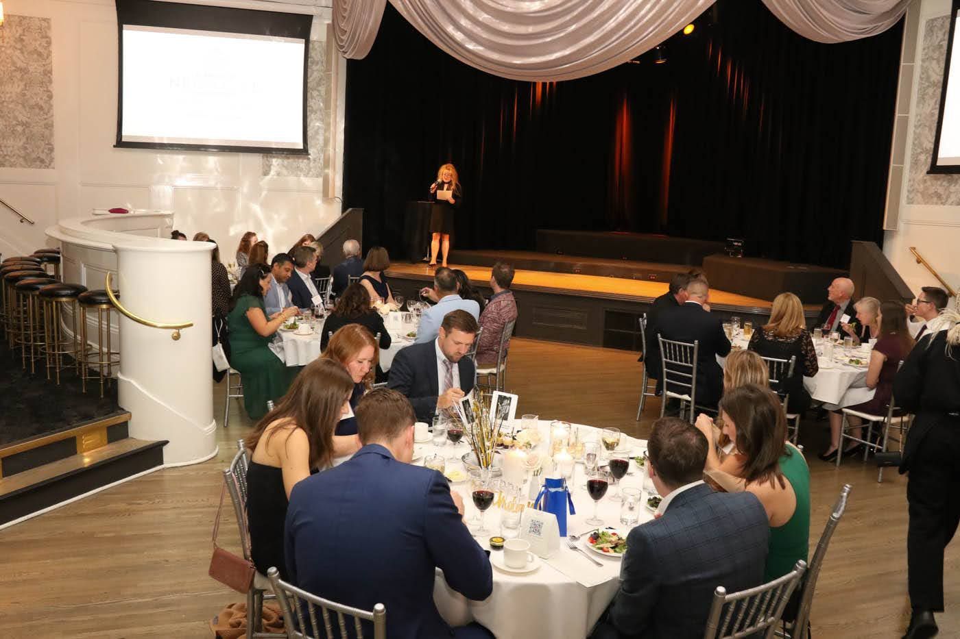 People seated at tables in a ballroom, a speaker on stage.