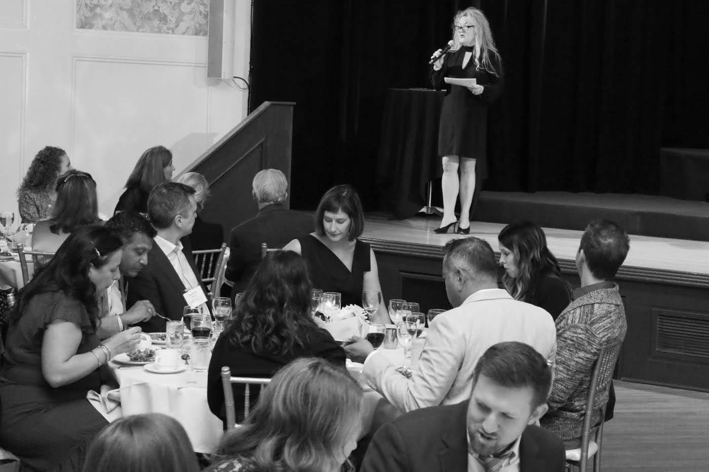 Woman speaking at an event, standing on a stage. People seated at tables in the foreground.