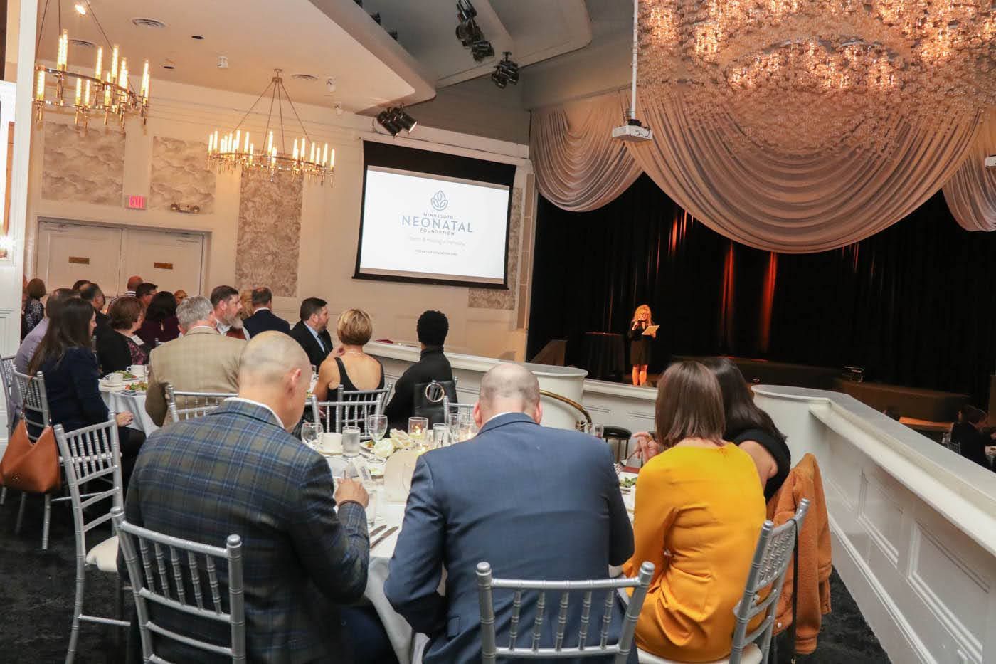 People seated at tables in a decorated event space, watching a presentation on stage.