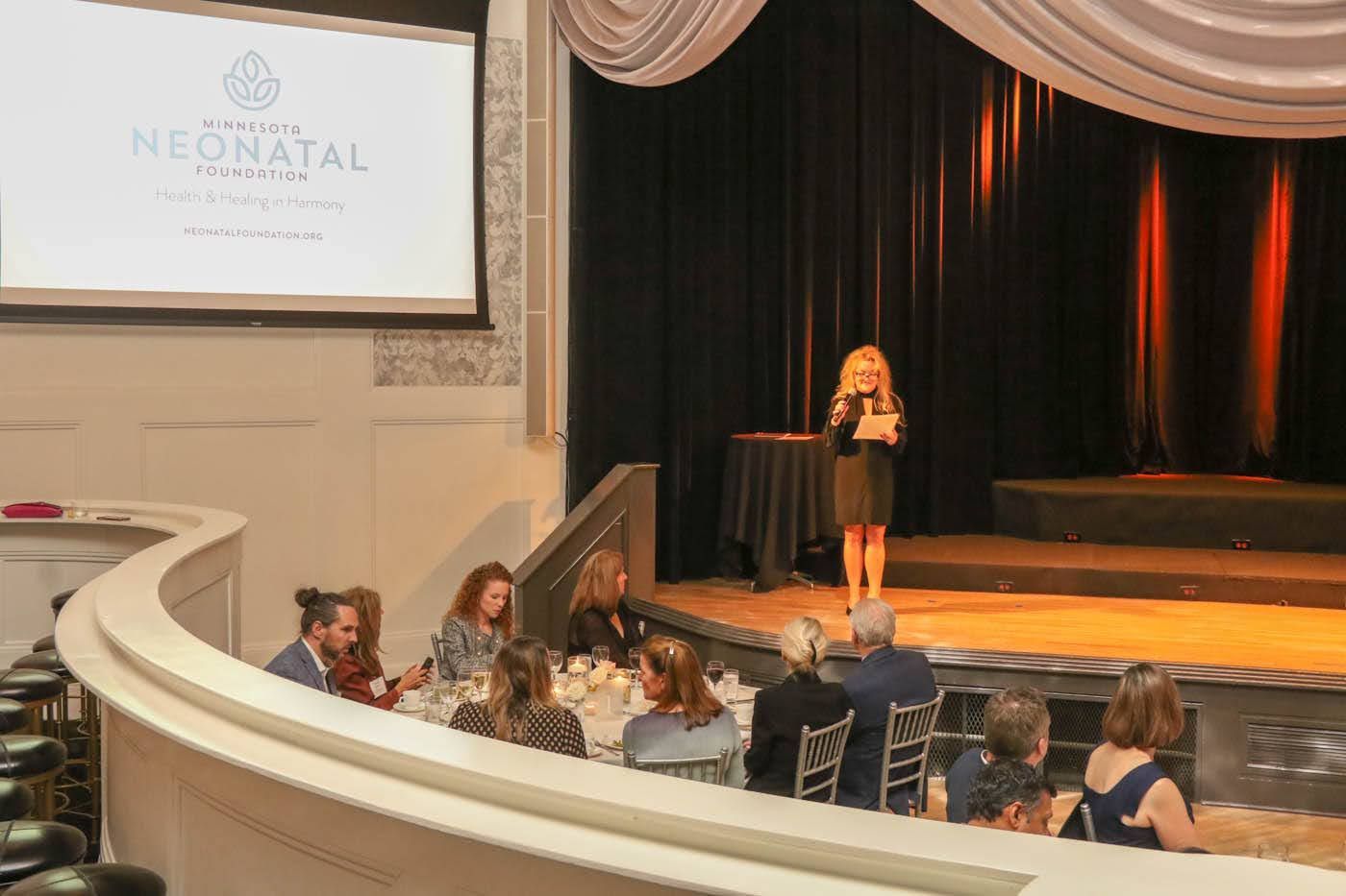 Woman speaking at a neonatal event, onstage. Attendees seated at tables in a theater setting.