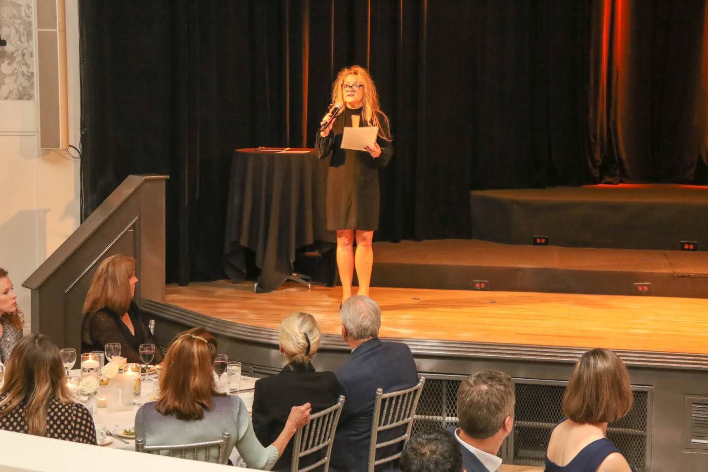 Woman speaking at a podium on a stage during an event. People seated at tables listen.