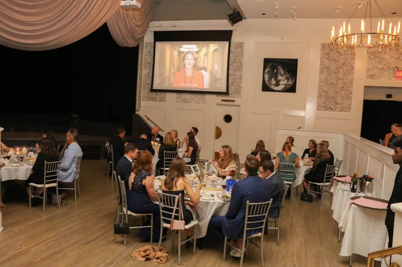 People seated at tables in a brightly lit banquet hall. A screen displays a video, and a chandelier hangs from the ceiling.