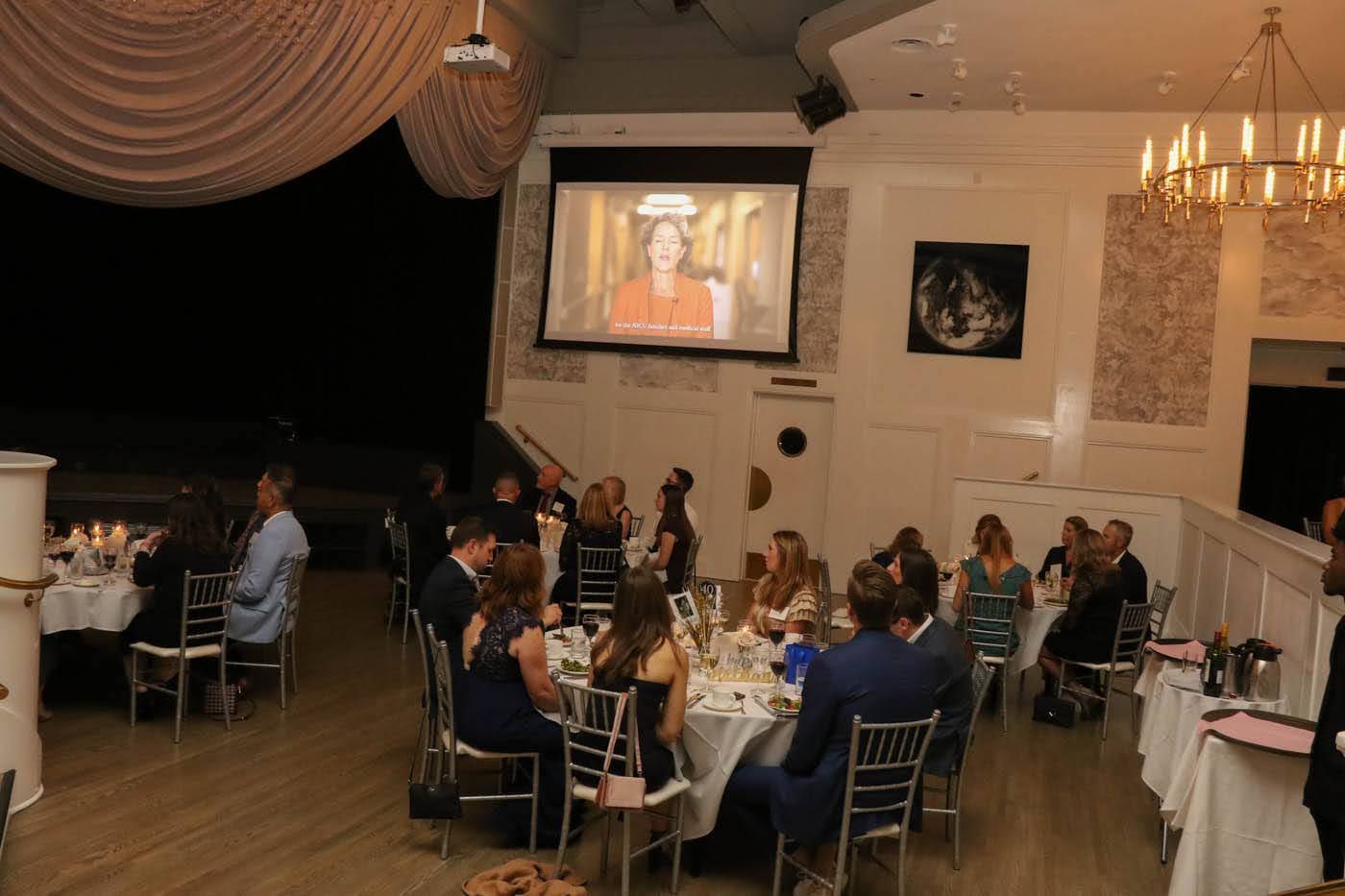 People seated at tables in a room watching a video on a screen. Elegant setting with chandeliers.