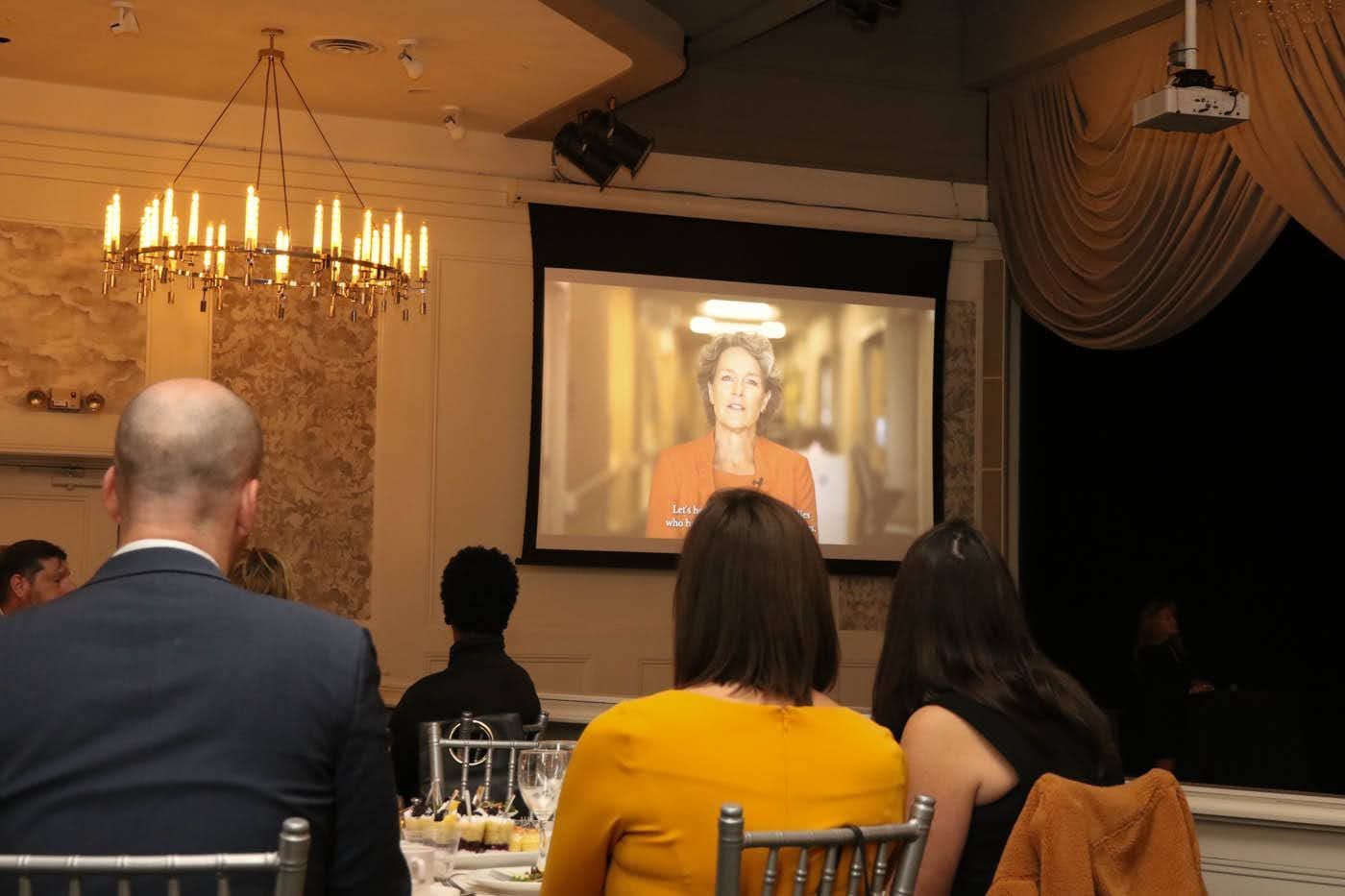 People watch a video on a screen. A woman speaks on the screen, and the audience sits at tables under a chandelier.