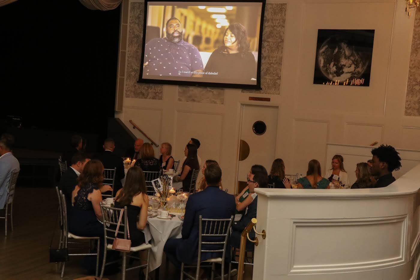 People seated at tables in a room watching a video on a screen.