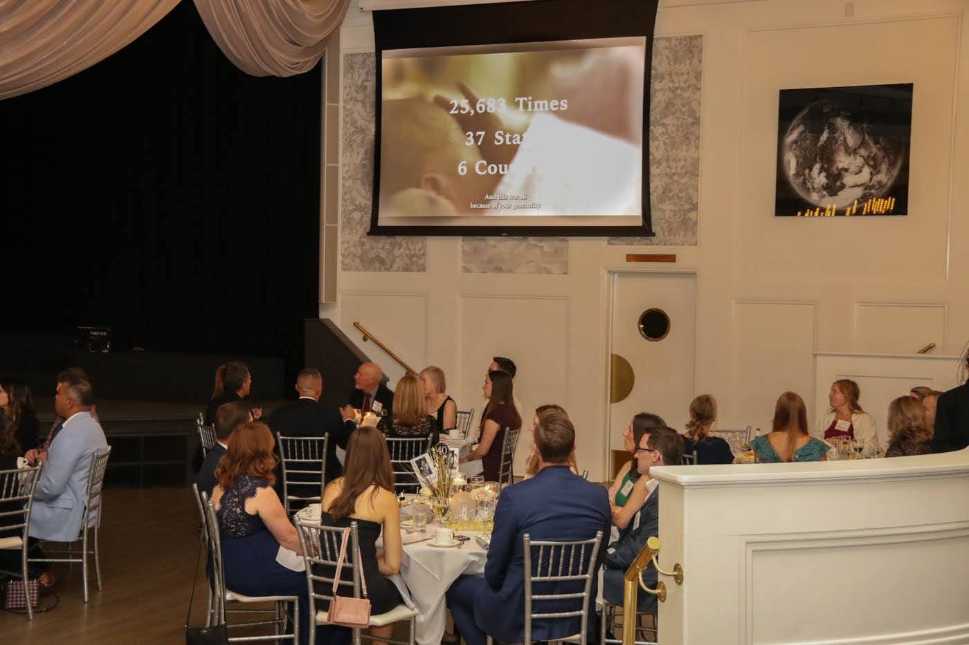 People seated at tables in a banquet hall, watching a screen with text.