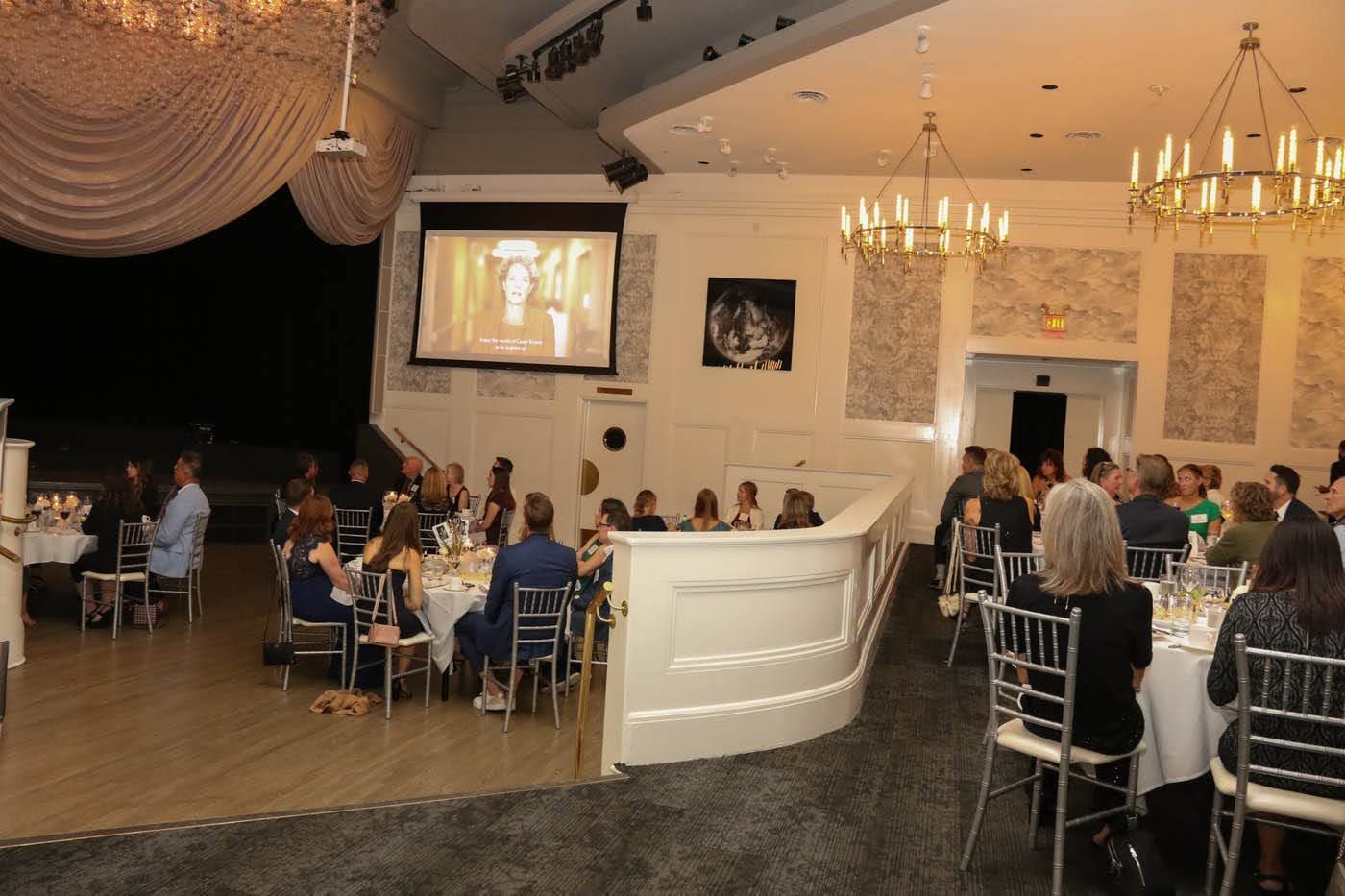 Elegant event venue with guests seated at tables, a stage, and a projected screen. Chandeliers hang above.