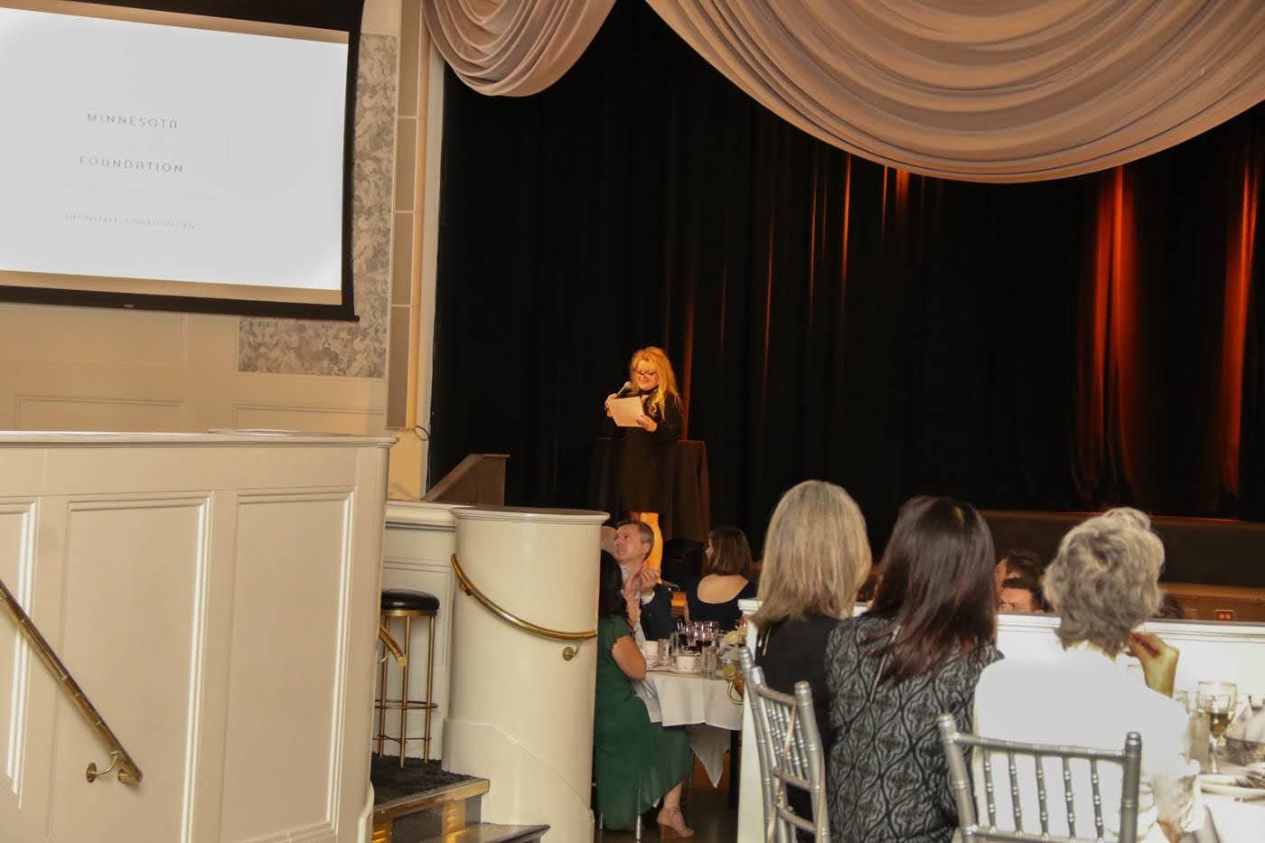 Woman speaking on a stage with a screen, audience seated at tables in a decorated room.