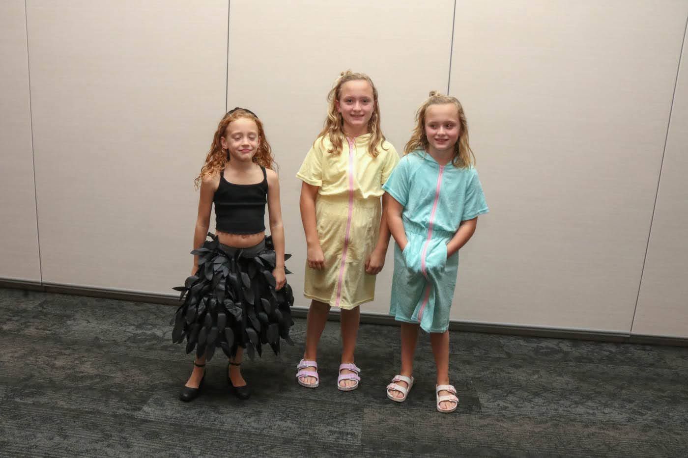 Three young girls in handmade dresses pose in front of a neutral backdrop.