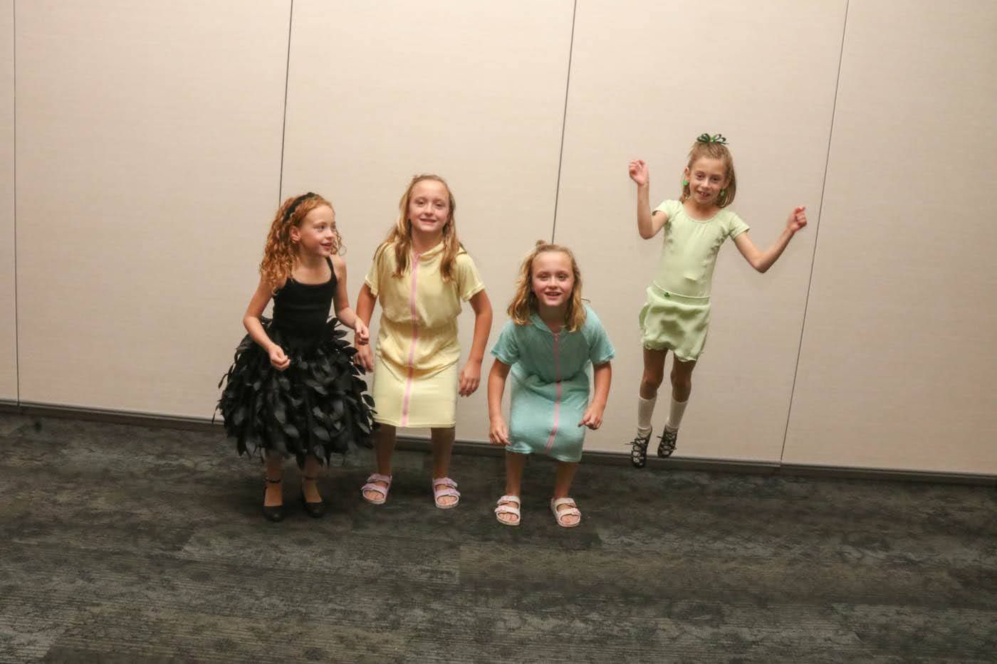 Four young girls jumping in a room with light-colored walls and dark carpet. Each wears a dress in black, yellow, teal, and lime green.