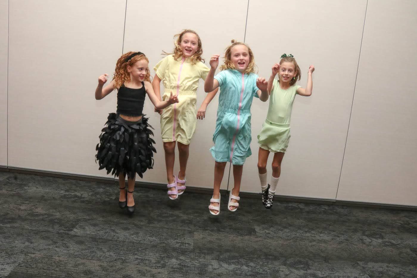 Four girls in various outfits jumping in front of a wall.