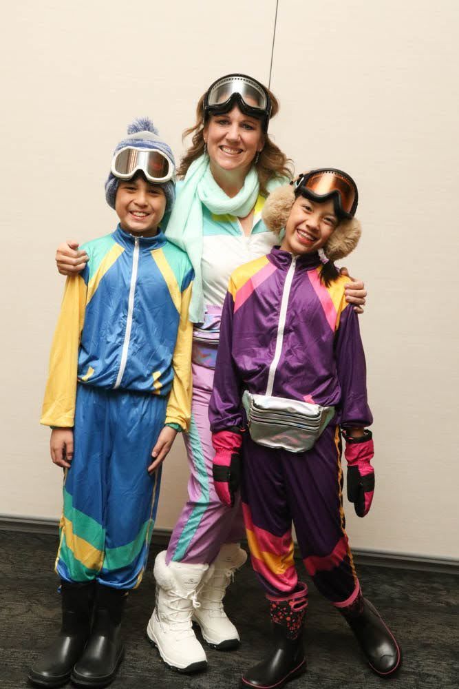 Three people in colorful vintage ski suits and goggles, standing indoors.