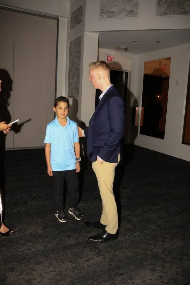 Boy in light blue shirt and dark pants stands beside a man in a navy blazer and tan pants.