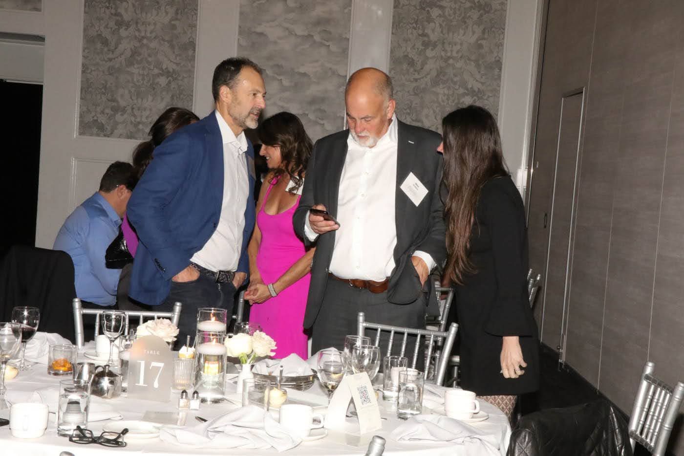 People at a formal event stand near a decorated table. Man in blazer looks at phone, others converse.