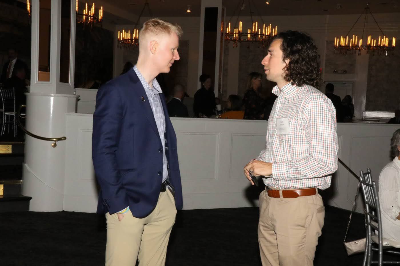 Two men converse in a dimly lit event space. One in a blue blazer and khakis, the other in a patterned shirt and tan pants.