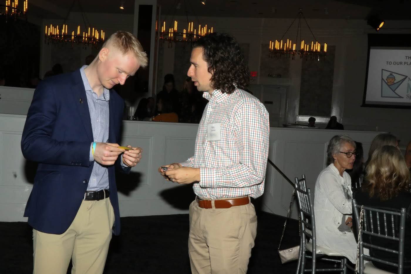 Two men exchange small items, indoors; one in blue blazer, one in striped shirt.