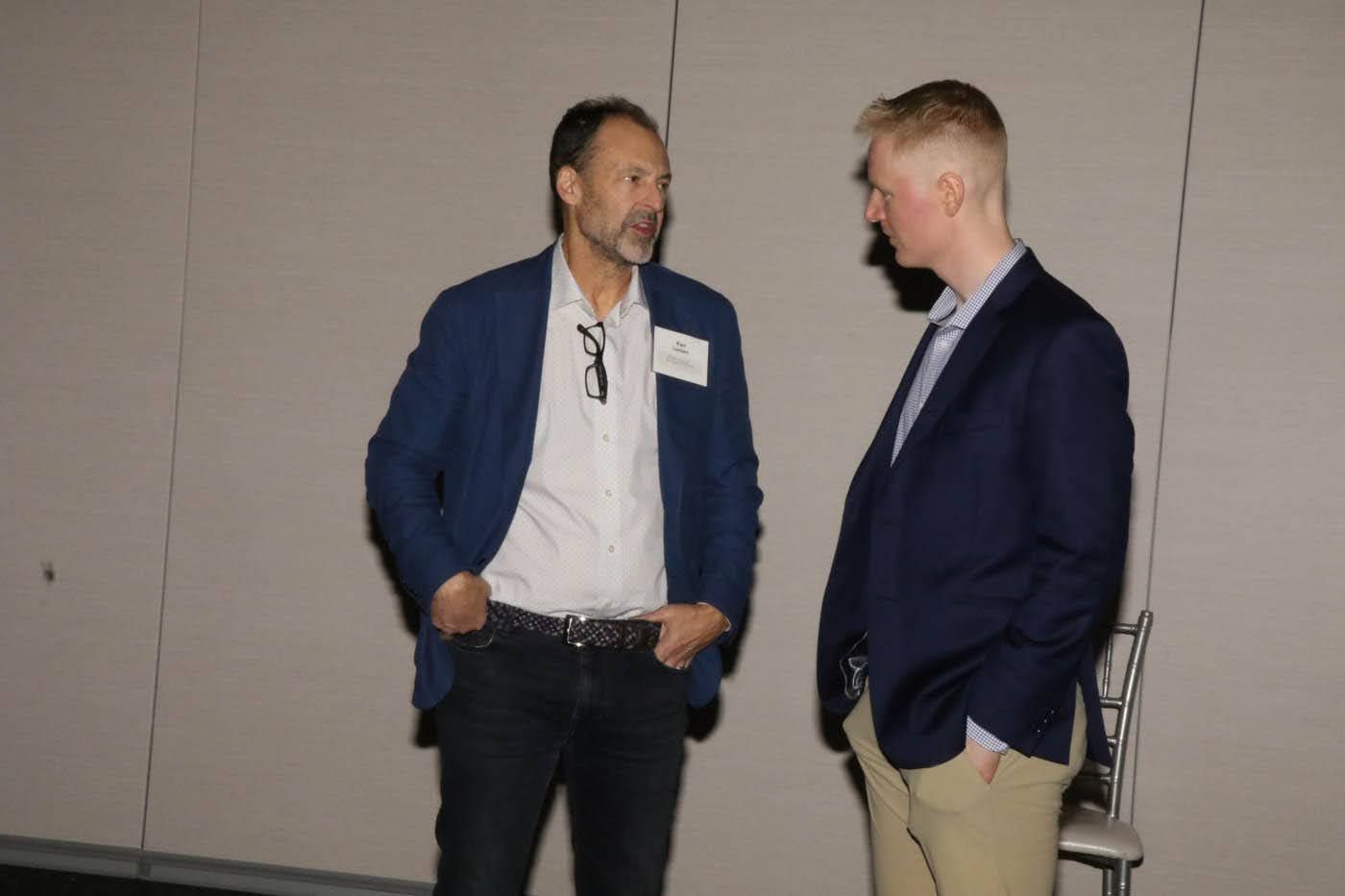 Two men in jackets conversing. One has hands in pockets, a name tag, the other stands next to him. Light-colored wall.