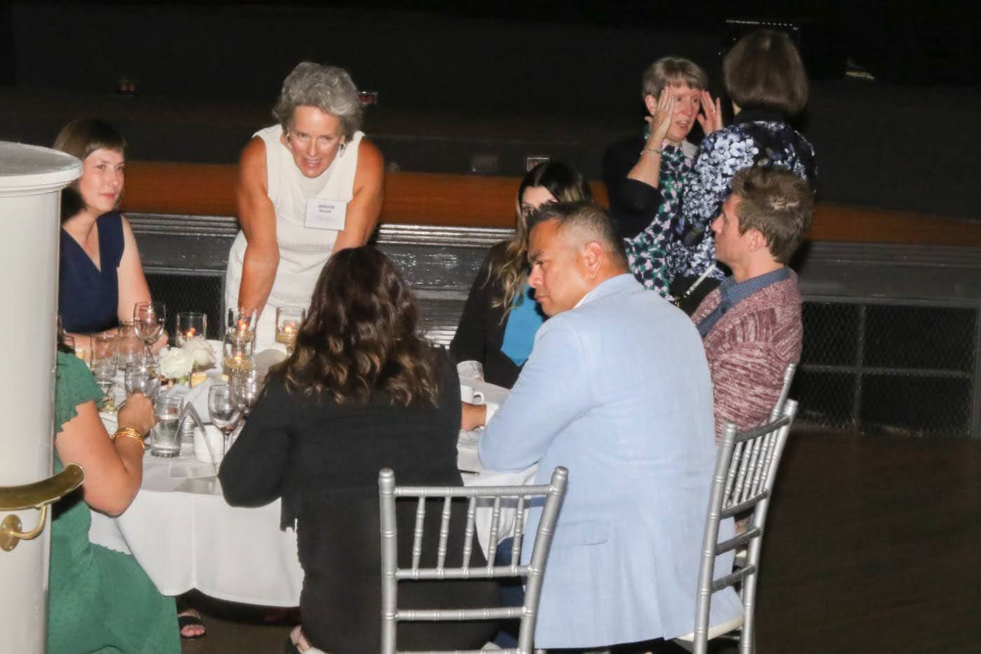 People at a round table, some looking at food, others talking. White tablecloth, silver chairs. Dark background.