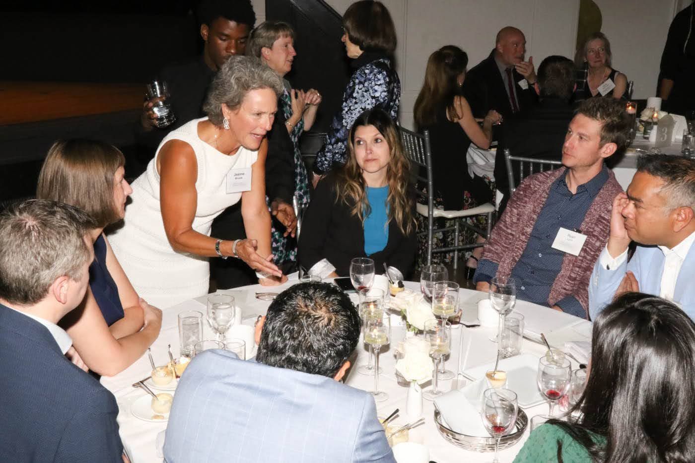 People at a dinner party, gathered around a table. A woman gestures with her hands as she speaks to the guests.
