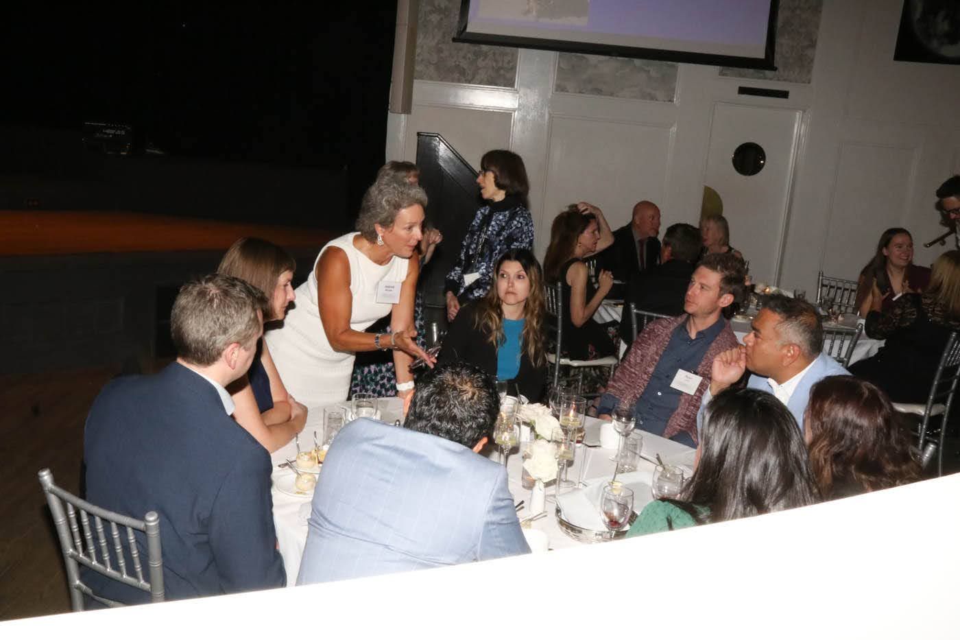 People at a dinner party, a woman in a white dress speaks animatedly to a group seated at a table.