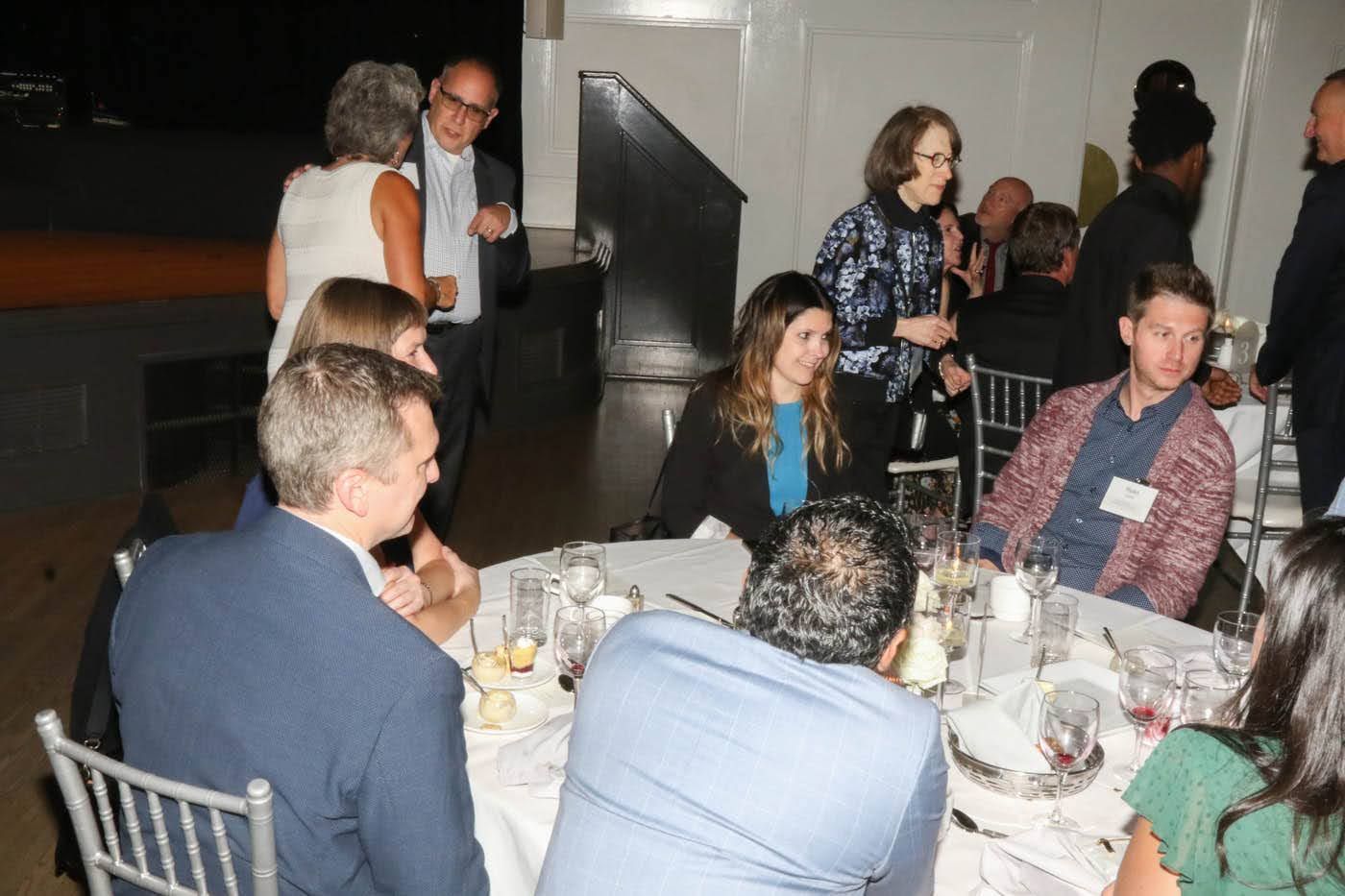 People at a formal event, seated at tables and socializing. Some are standing and talking.
