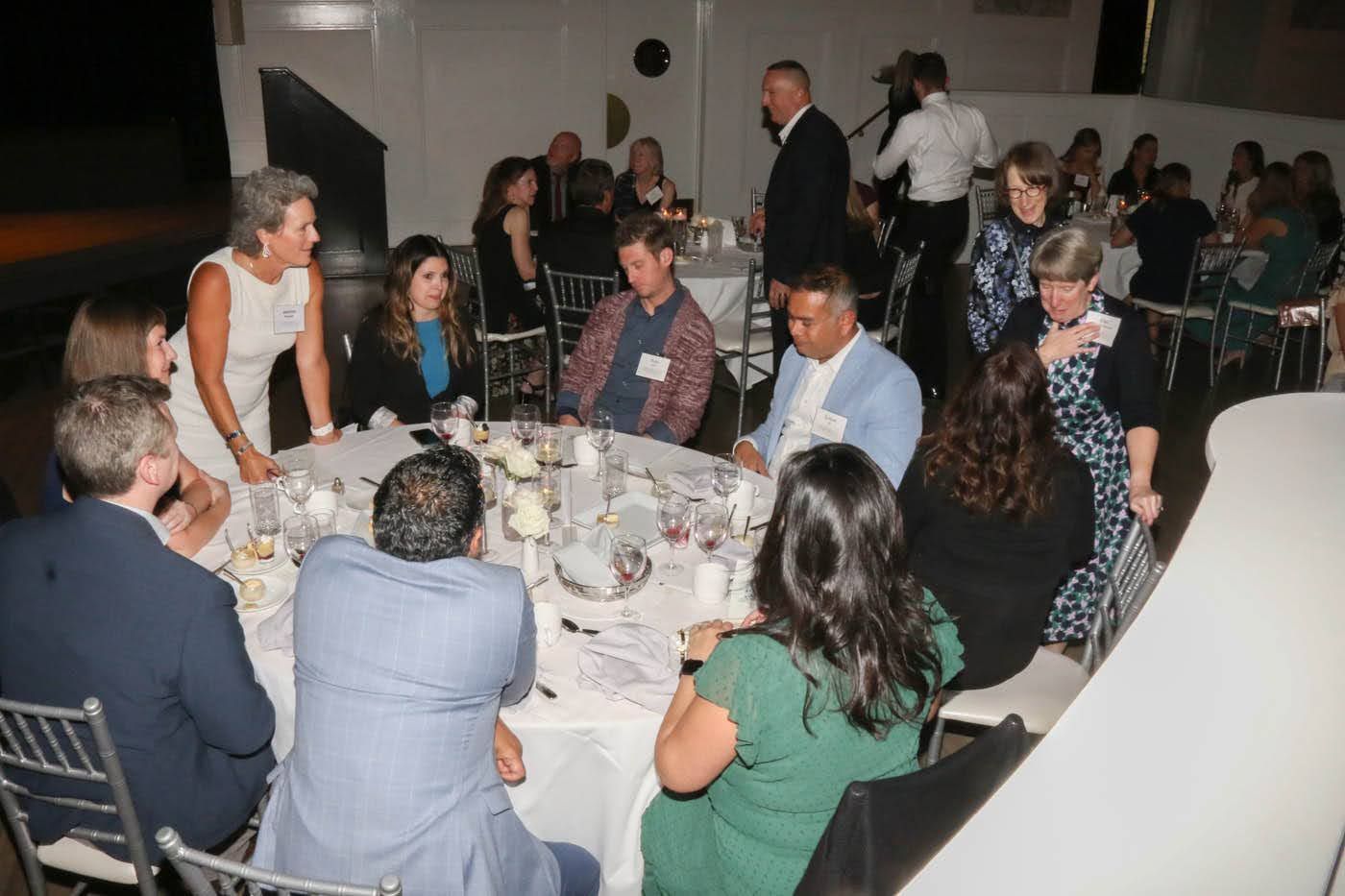 People at tables in a brightly lit room with white tablecloths. A woman in a white dress adjusts a table setting.