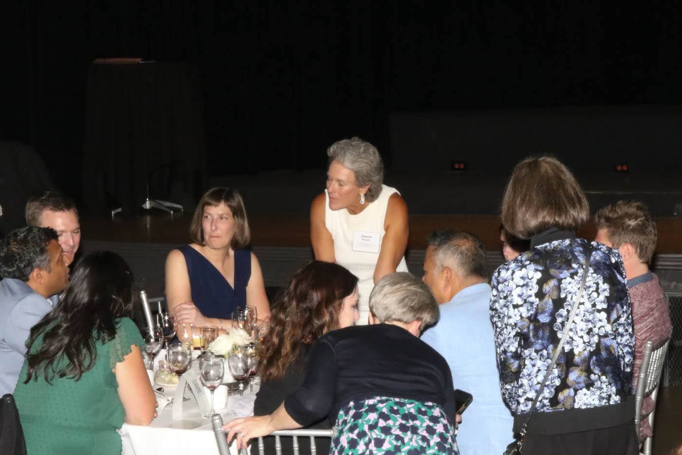 People seated at a table, conversing. One woman stands addressing the group. Evening event, dim lighting.