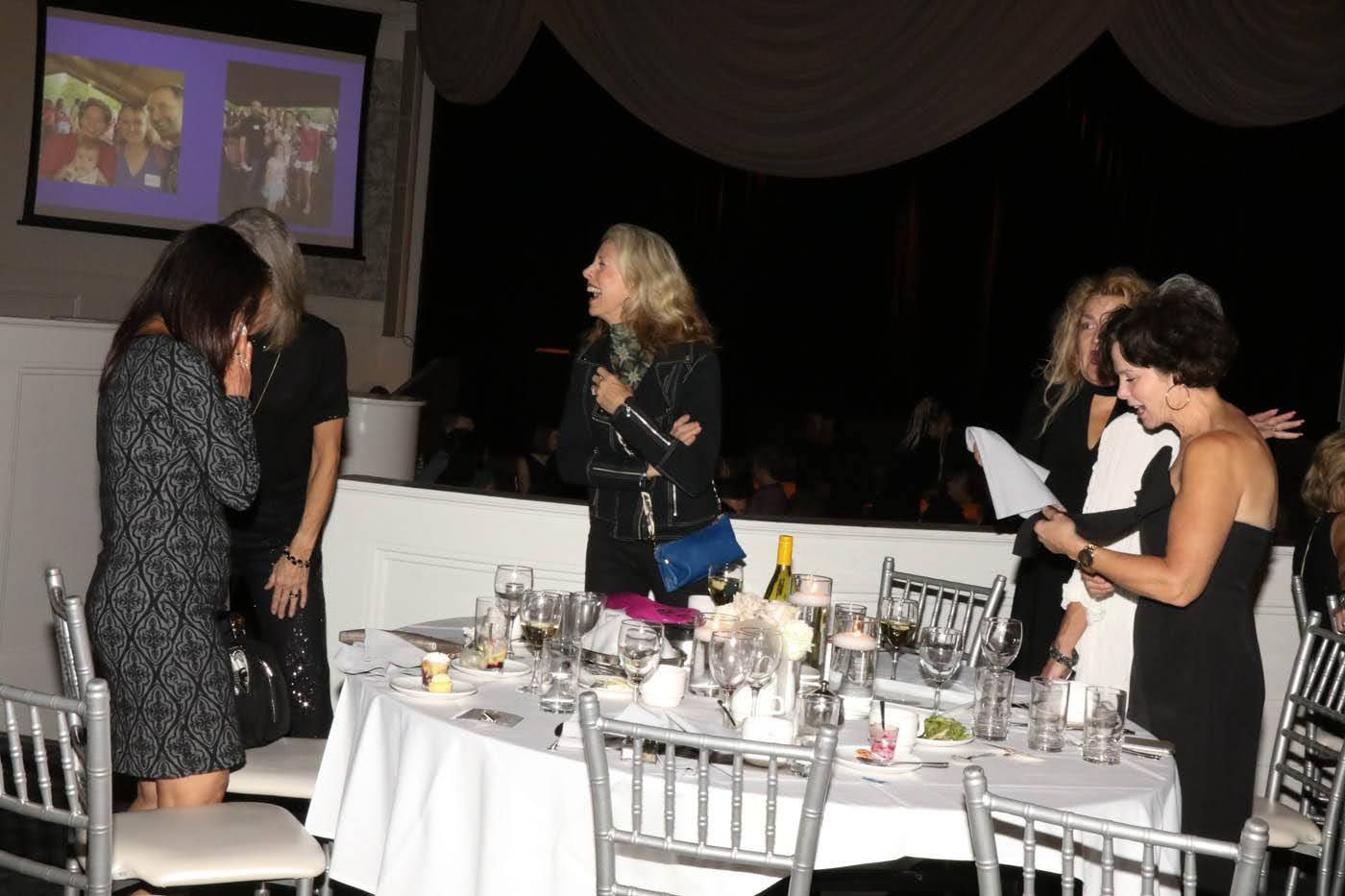 Guests gather at a formal event, tables set with silverware, screen in background, one woman reads from paper.