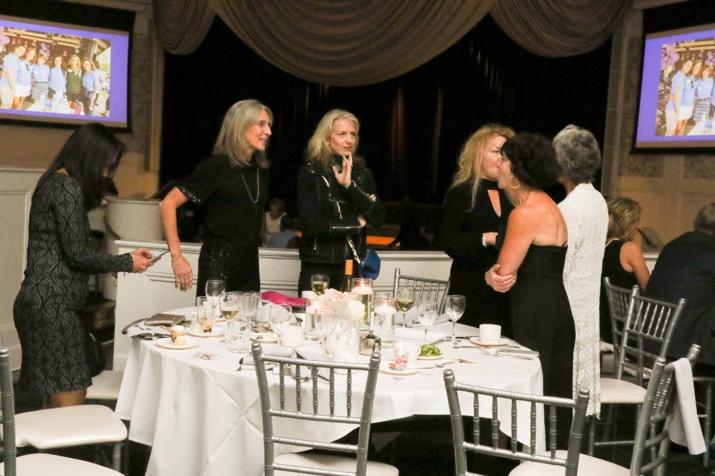 People at an event. Some are at a round table, others are talking. Two screens show a slideshow.