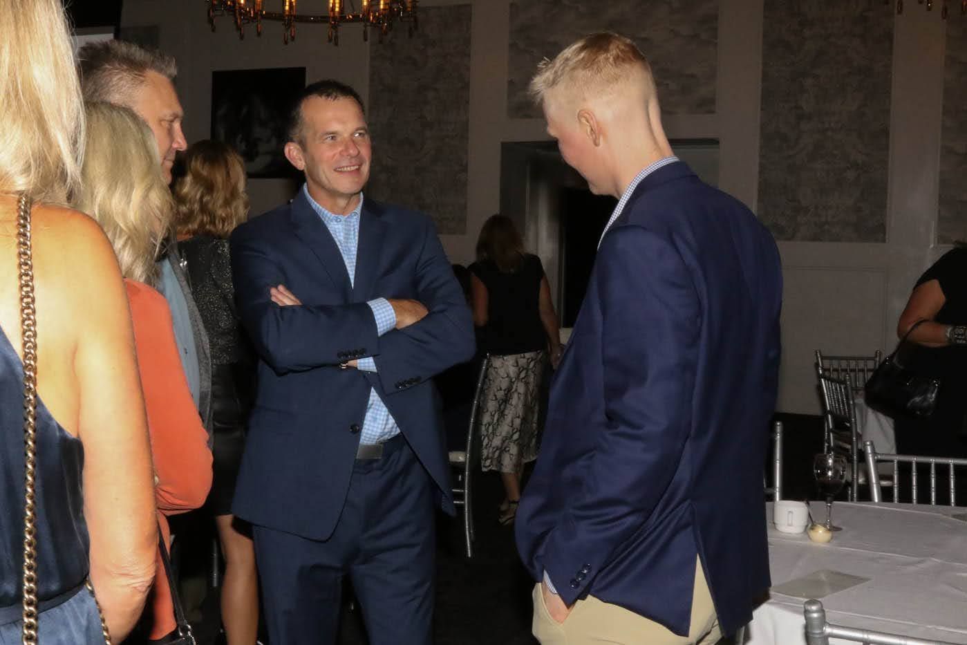 Two men in suits converse at an event, surrounded by other guests in a room.