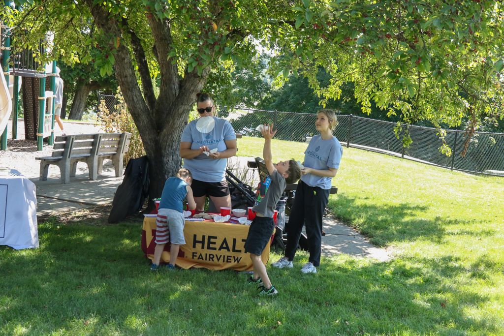 People at a HealthFair table under a tree. One person tosses something up, others look on. Sunny day.