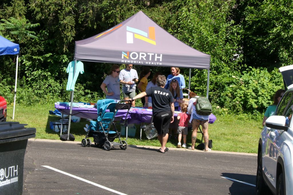 People at a booth with a gray canopy. Staffer gestures, a stroller, trees, and a parked car are present.