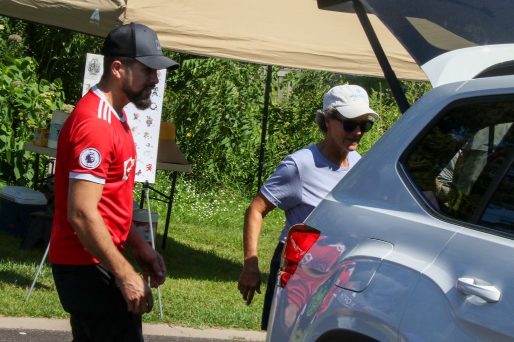 Man in red jersey and woman by car, under tent, outside.